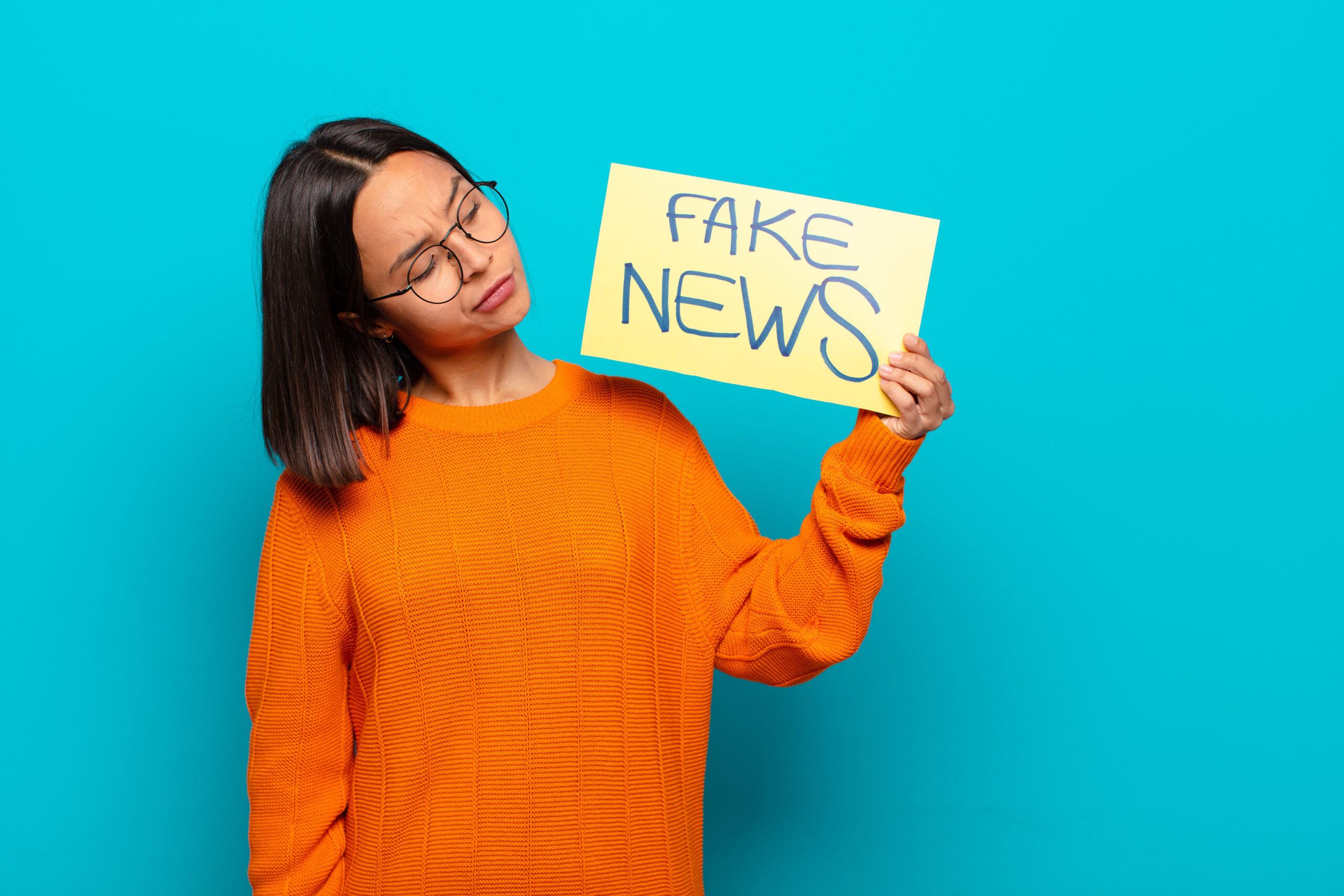 A woman in an orange sweater holds a sign reading "Fake News" against a blue background.