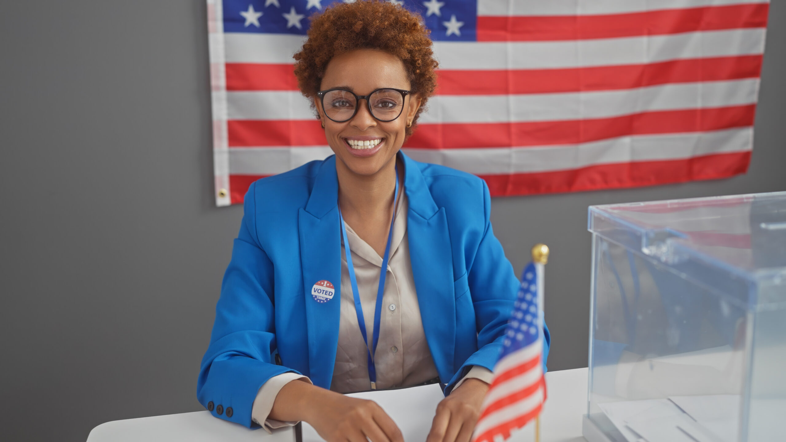 A smiling african american woman with a 'voted' sticker, positioned before an american flag inside a polling place.